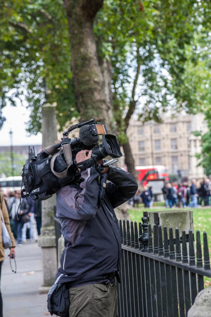 Cameraman professionnel filme avec sa caméra sur l'épaule en extérieur. Foule et bâtiment flous en arrière-plan.