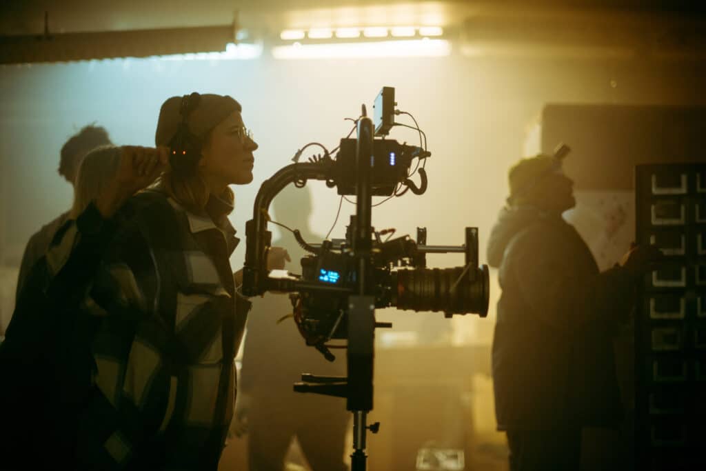 Personne regarde dans une caméra de tournage professionnelle sur un plateau. Autres membres de l'équipe visibles dans la brume lumineuse.