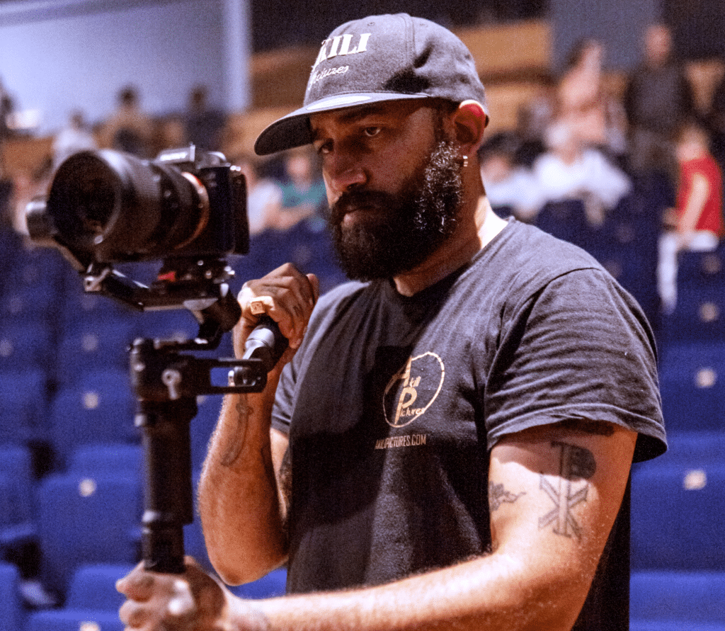 Vidéaste professionnel Akili Pictures filme un événement Cameraman concentré, casquette AKILI Pictures, filme avec caméra sur gimbal. T-shirt AKILI Pictures.