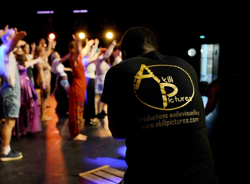 Homme de dos, t-shirt noir Akili Pictures, observe des danseurs sur scène colorée. Texte: productions audiovisuelles, www.akilipictures.com.